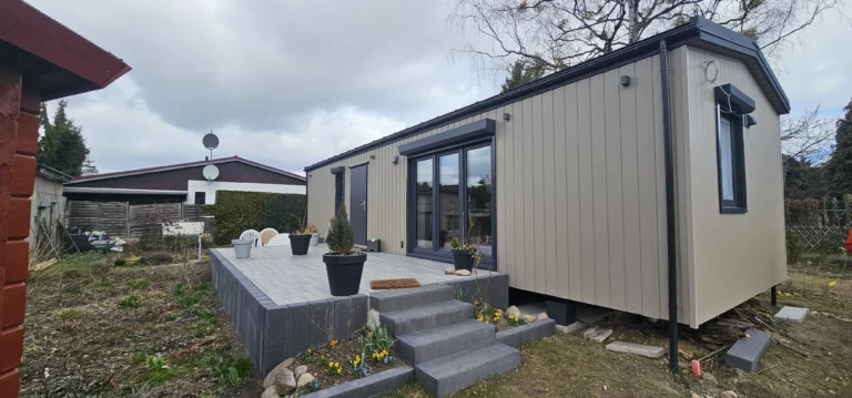 Perspektivische Ansicht eines winterfesten Compact Hauses der MONO-Serie mit hellbeiger Holzfassade, großer Holzterrasse, grauen Blockstufen aus Beton und bepflanzten Blumentöpfen.