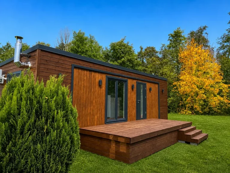 Individuell angefertigte Holzterrasse mit Treppenstufen an einem modernen Mobilheim von Compact Haus auf einer gepflegten Rasenflaeche vor einer Waldkulisse.