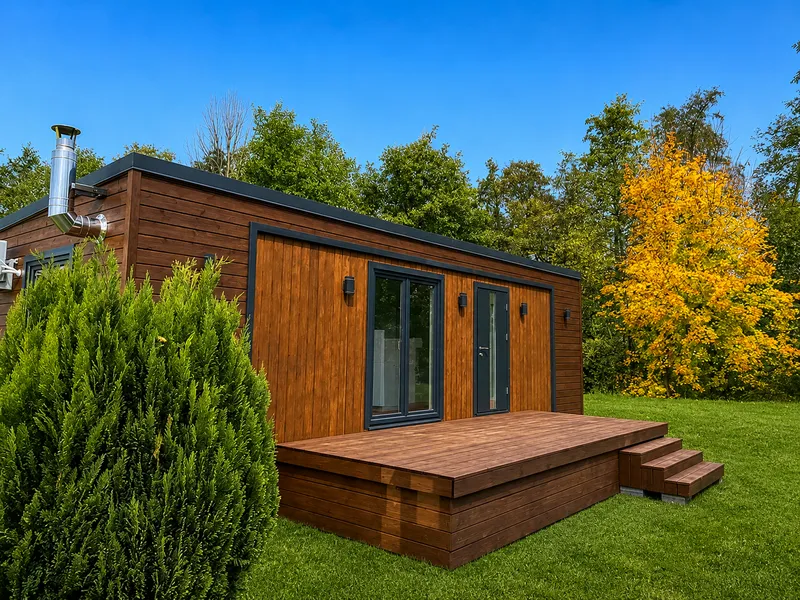 Individuell angefertigte Holzterrasse mit Treppenstufen an einem modernen Mobilheim von Compact Haus auf einer gepflegten Rasenflaeche vor einer Waldkulisse.
