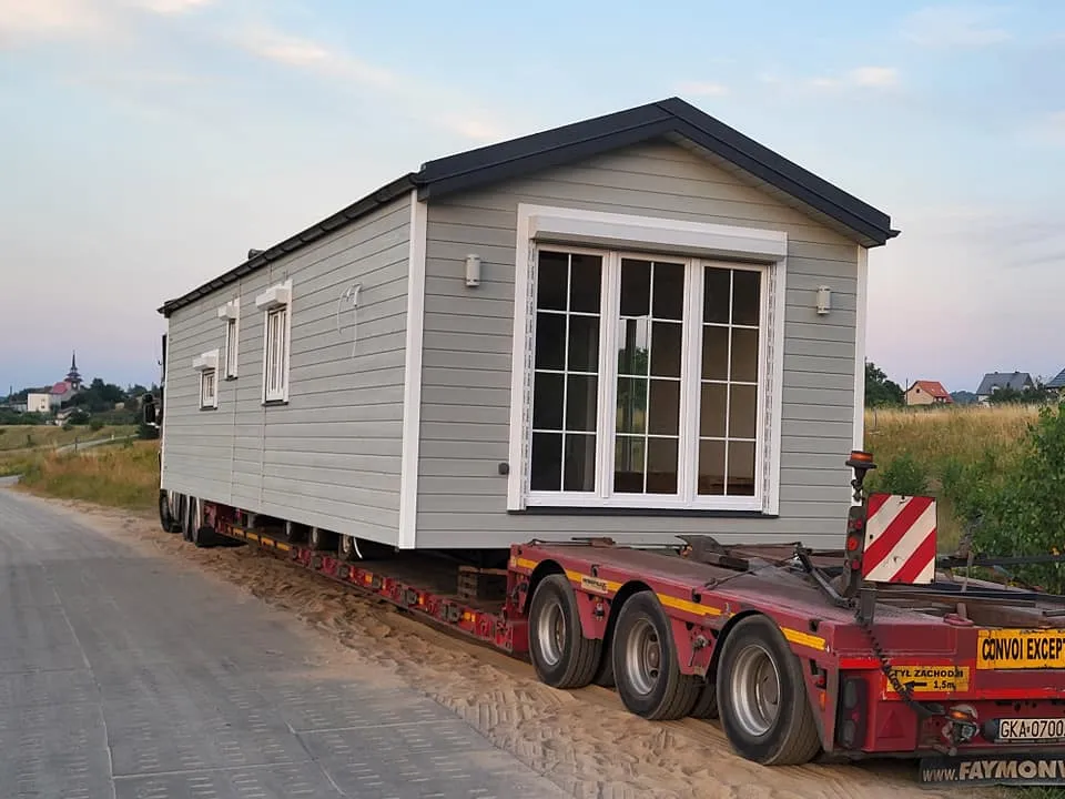 Spezialtransport eines winterfesten Mobilheims der MONO-Serie von Compact Haus mit hellgrauer Holzfassade auf einem roten Schwerlast-Tieflader.