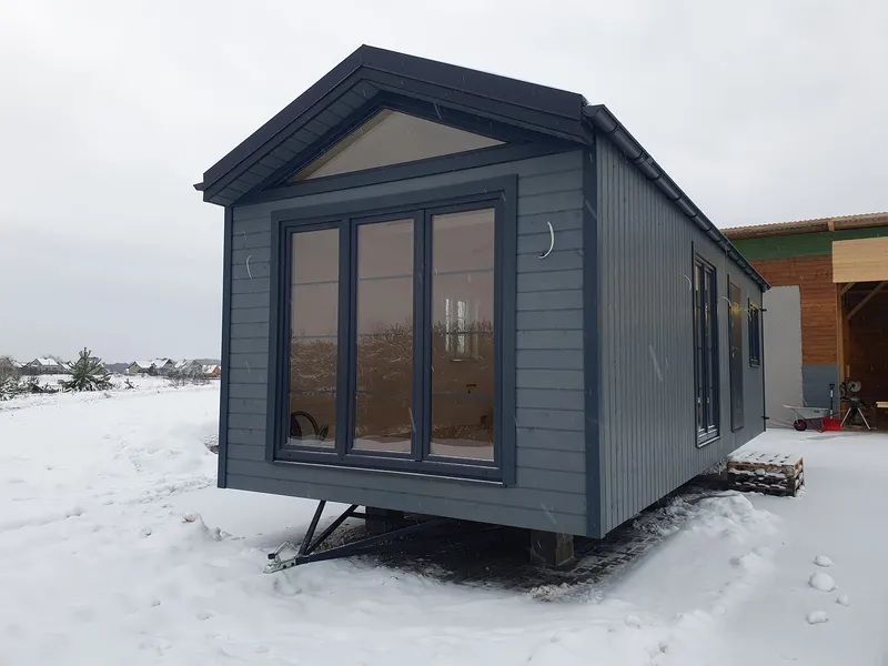 Winterfestes Mobilheim von Compact Haus in Grau mit großen Giebelfenstern und sichtbarem Trailer-Fahrgestell.