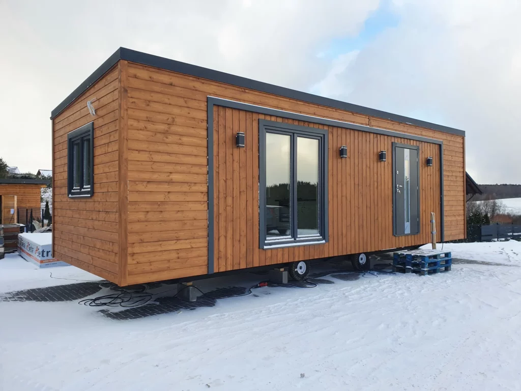 Winterfestes Mobilheim der MONO-Serie von Compact Haus mit brauner Holzfassade, anthrazitfarbenen Fenster- und Türelementen auf einem schneebedeckten Grundstück.