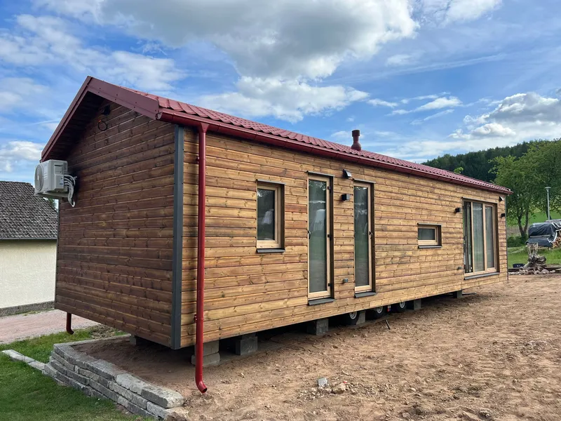 Winterfestes Mobilheim der MONO-Serie von Compact Haus in Holzständerbauweise mit natürlicher Echtholzfassade, rotem Satteldach, bodentiefen Fenstern und großer Glasschiebetür.