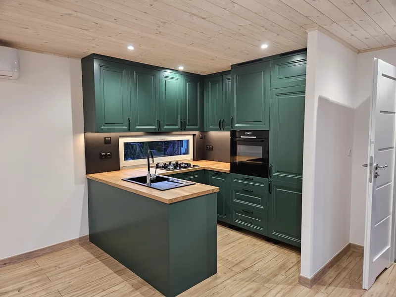 Großzügige Einbauküche in U-Form mit waldgrünen Fronten im Landhausstil, integrierter Küchentheke, Gasherd und hölzernem Fußboden in einem Tiny House der MONO-Serie.