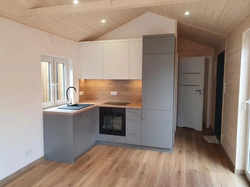 Moderne zweifarbige L-Küche in einem Tiny House der MONO-Serie von Compact Haus mit grauen Unterschränken, weißen Oberschränken und einer Rückwand in Holzoptik.