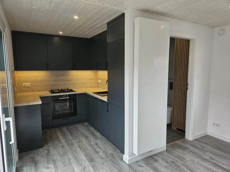 Moderne anthrazitfarbene U-Küche im Landhausstil mit heller Holzarbeitsplatte, integriertem Backofen und einer weißen Schiebetür im Innenraum eines Tiny Houses der MONO-Serie.