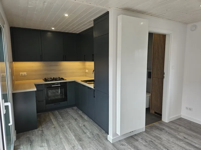 Moderne anthrazitfarbene U-Küche im Landhausstil mit heller Holzarbeitsplatte, integriertem Backofen und einer weißen Schiebetür im Innenraum eines Tiny Houses der MONO-Serie.