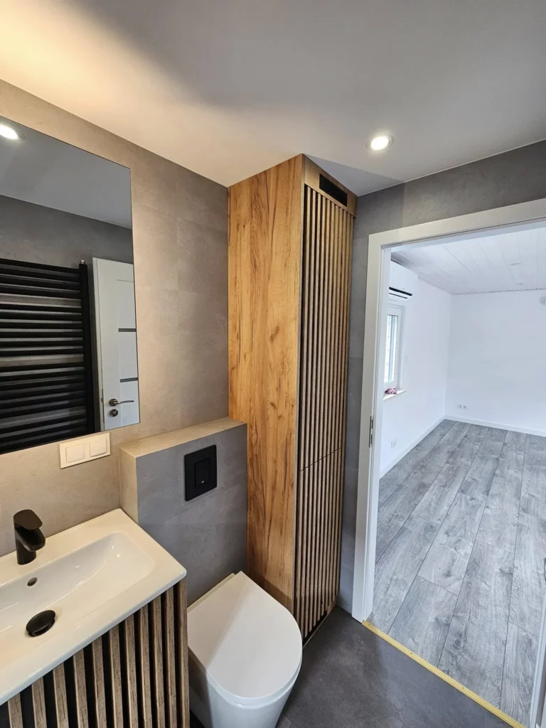Badezimmer in einem Compact Haus der MONO-Serie mit maßgefertigtem Hochschrank aus Holzlamellen, Wand-WC, Spiegel und Blick durch die geöffnete Tür in das angrenzende Zimmer mit grauem Vinylboden.