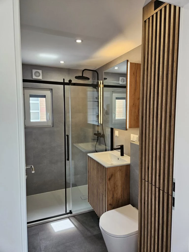 Elegantes Badezimmer im Mobilheim der MONO-Serie von Compact Haus mit bodengleicher Glasdusche, Waschtischunterschrank aus Holzlamellen, beleuchtetem Spiegelschrank und moderner Wandverkleidung in Betonoptik.