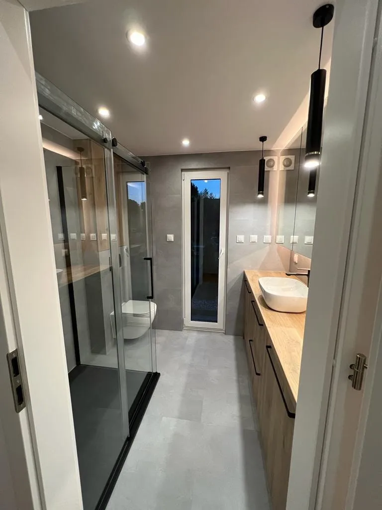 Elegantes Badezimmer im Barnhouse-Mobilheim der MONO-Serie von Compact Haus mit anthrazitfarbener Glasdusche, langem Holz-Waschtisch, Aufsatzwaschbecken und modernen Pendelleuchten.