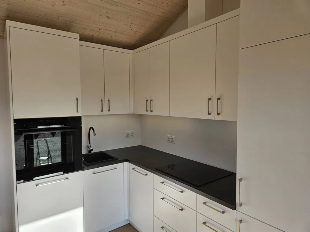 Moderne weiße Einbauküche in L-Form mit dunkler Arbeitsplatte, integriertem Backofen und flächenbündigem Kochfeld unter einer hellen Holz-Giebeldecke im Compact Haus der MONO-Serie.