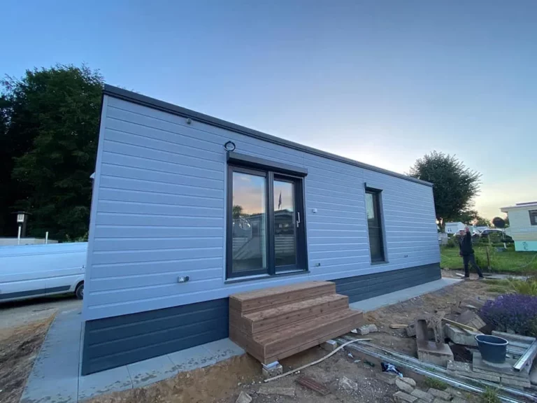Modernes winterfestes Mobilheim der MONO-Serie von Compact Haus mit hellgrauer Außenfassade, anthrazitfarbenem Sockel und einer hölzernen Eingangstreppe.