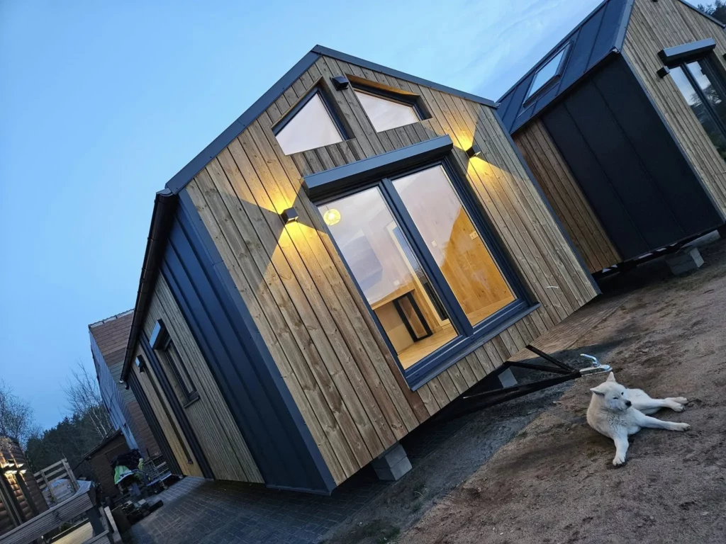 Modernes winterfestes Mobilheim der MONO-Serie von Compact Haus mit vertikaler Holzfassade, anthrazitfarbenen Metallelementen, großen Giebelfenstern und stimmungsvoller Außenbeleuchtung in der Dämmerung.